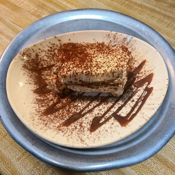A slice of tiramisu dusted with cocoa powder and drizzled with chocolate sauce on a white plate.