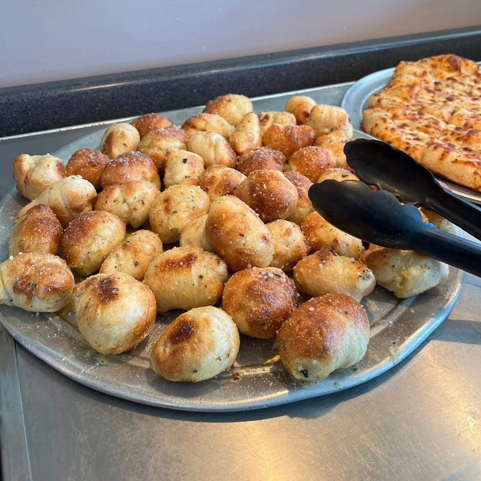 A metal tray filled with golden-brown garlic knots, sprinkled with herbs and cheese, next to a cheese pizza.