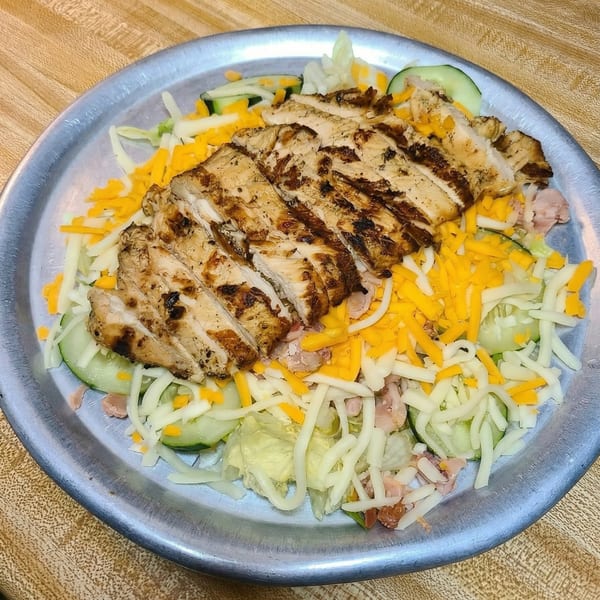 Grilled chicken salad with shredded cheese, cucumbers, and lettuce on a metal plate.
