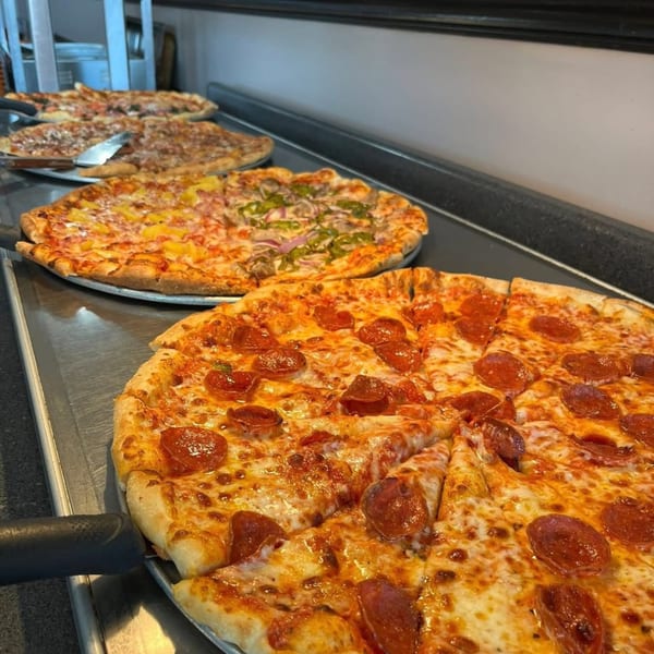 Multiple large pizzas with various toppings, including pepperoni, pineapple, and vegetables, on a metal counter.