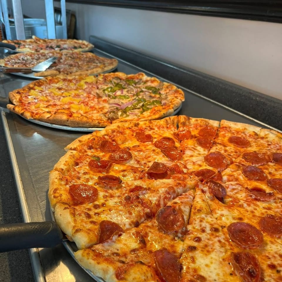 Multiple large pizzas with various toppings, including pepperoni, pineapple, and vegetables, on a metal counter.